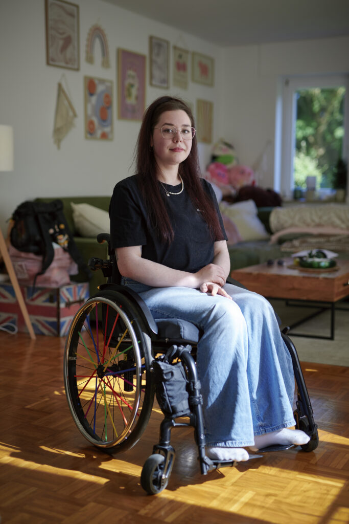 Eine junge Frau schaut mit ernsten Blick in die Kamera. Sie hat lange braune Haare und trägt Jeans und ein schwarzes T-Shirt. Sie sitz in einem Rollstuhl. Im Hintergrund ist ein Wohnzimmer unscharf zu sehen.