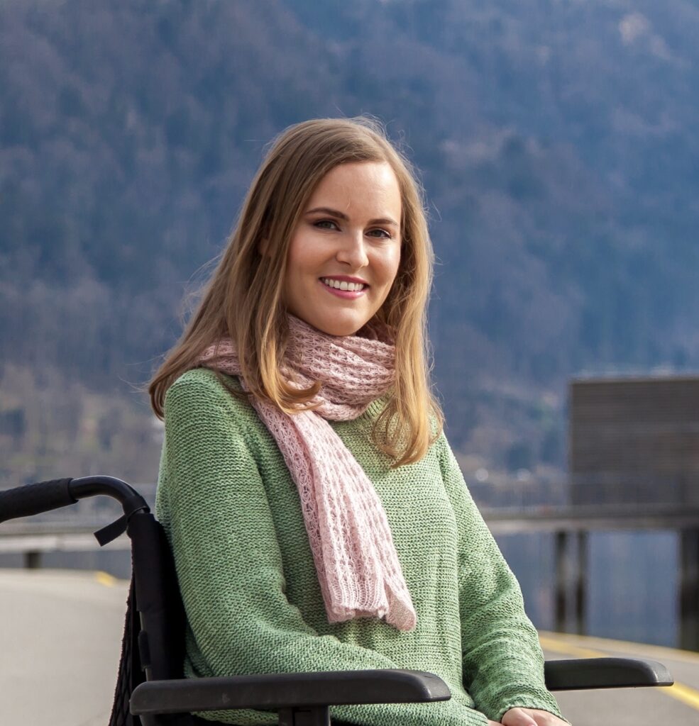Eine Frau schaut mit freundlichen Blick in die Kamera. Sie hat dunkelblonde schulterlange Haare und trägt einen rosa Schal und einen grünen Pullover. Sie sitzt in einem Rollstuhl. Im Hintergrund ist ein Berg und ein See unscharf zu sehen.