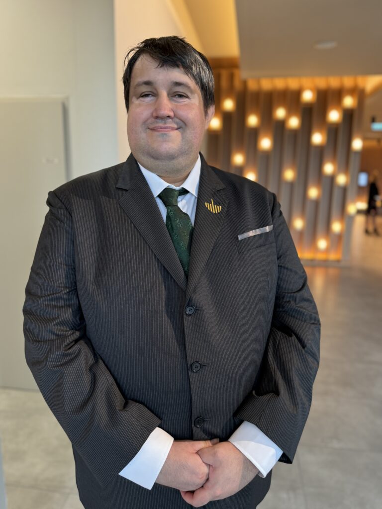 Ein Mann schaut mit freundlichen Blick in die Kamera. Er hat kurze schwarze Haare und trägt einen Anzug.