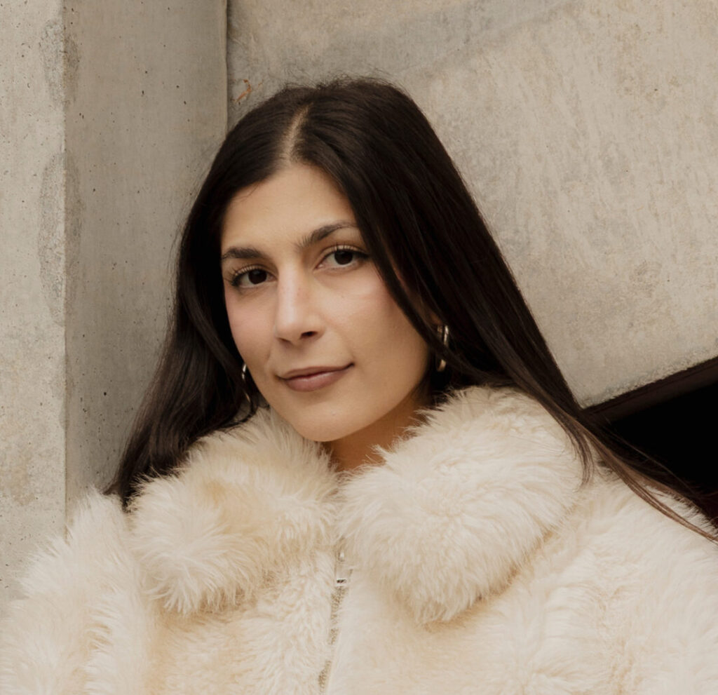 Eine junge Frau schaut mit leichten freundlichen Blick in die Kamera. Sie hat dunkelbraune glatte Haare und dunkelbraune Augen. Sie trägt eine weiße Felljacke.