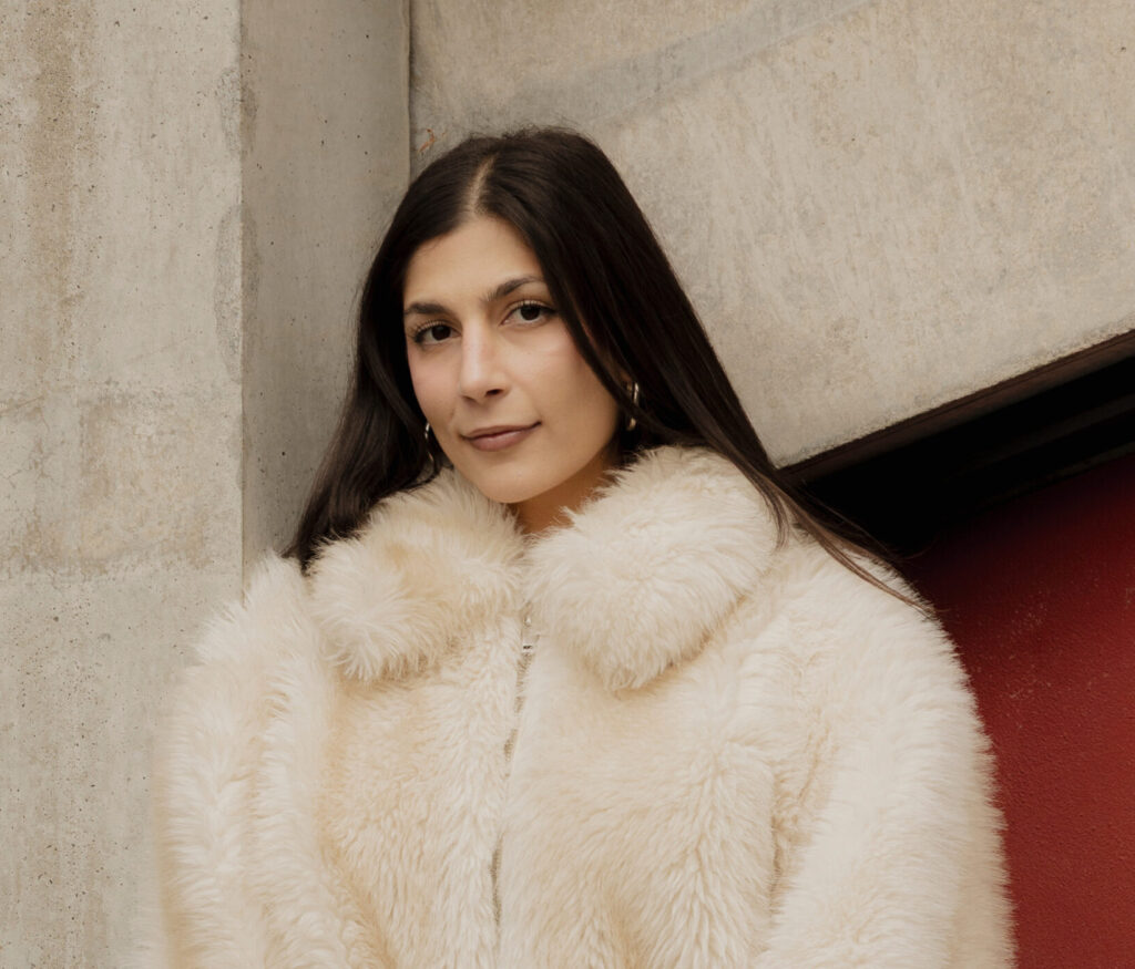 Eine junge Frau schaut mit leichten freundlichen Blick in die Kamera. Sie hat dunkelbraune glatte Haare und dunkelbraune Augen. Sie trägt eine weiße Felljacke.