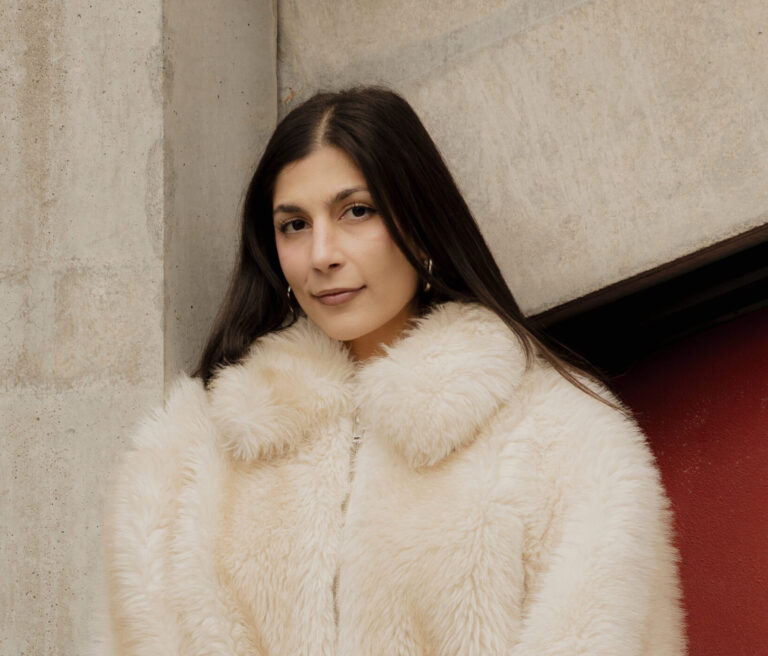 Eine junge Frau schaut mit leichten freundlichen Blick in die Kamera. Sie hat dunkelbraune glatte Haare und dunkelbraune Augen. Sie trägt eine weiße Felljacke.