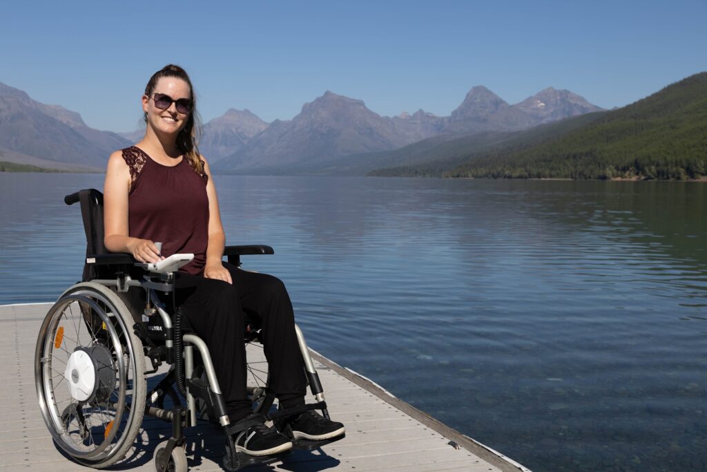 Eine Frau sitzt in einem Rollstuhl. Sie lächelt in die Kamera. Sie trägt eine Sonnenbrille, die Haare sind zusammengebunden. Im Hintergrund ist ein See und mehrere Berge.