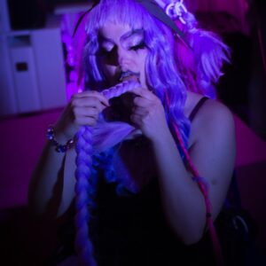 Eine Person mit lila Haare hält einen Zopf in de Hand. Sie trägt Makeup, glitzernden Liedschatten und hat künstliche Wimpern. Im Hintergrund sind Neon-Lichter zu sehen.