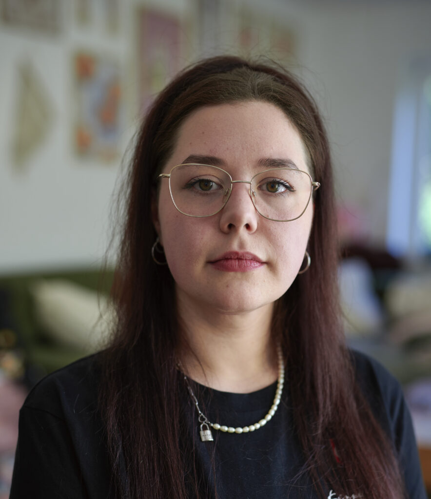 Eine junge Frau schaut mit ernstem Blick in die Kamera. Sie trägt eine Brille und hat lange braune Haare.
