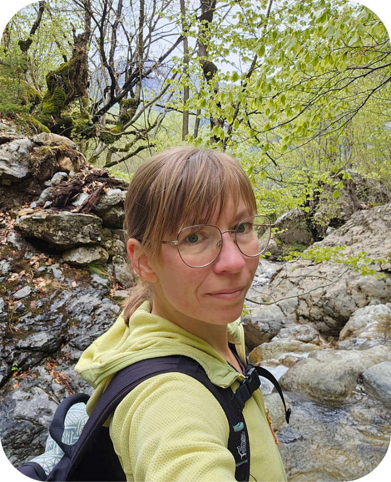Eine Frau schaut in die Kamera. Sie trägt eine Brille. Sie ist im Wald.