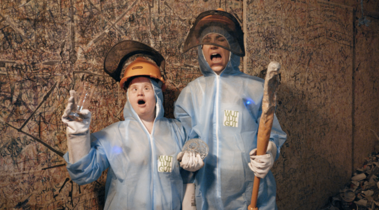 Das Bild zeigt zwei Frauen, die wütend schreien. Sie tragen einen blauen Schutzanzug und orange Helme. Die Frau rechts hält einen Hammer in der Hand. Die linke Frau ein Glas.