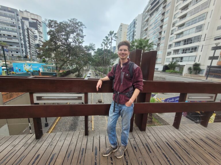 Ein junger Mann steht an ein Geländer gelehnt. Er trägt ein rotes Hemd und helle Jeans.