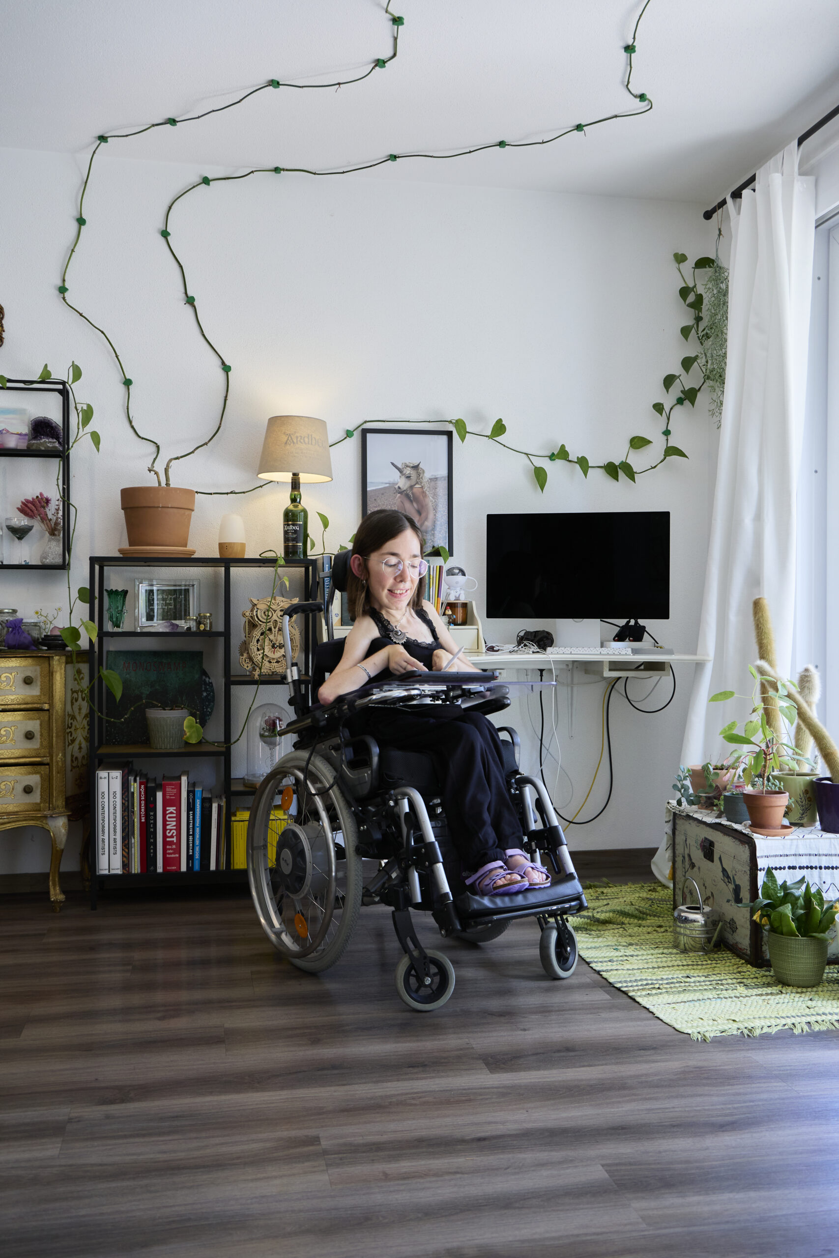 Eine Frau sitzt in einem Rollstuhl. Sie ist in einem Zimmer. Im Hintergrund ist ein weiße Wand, davor ein Computerbildschirm, eine Pflanze und ein Bücherregal.