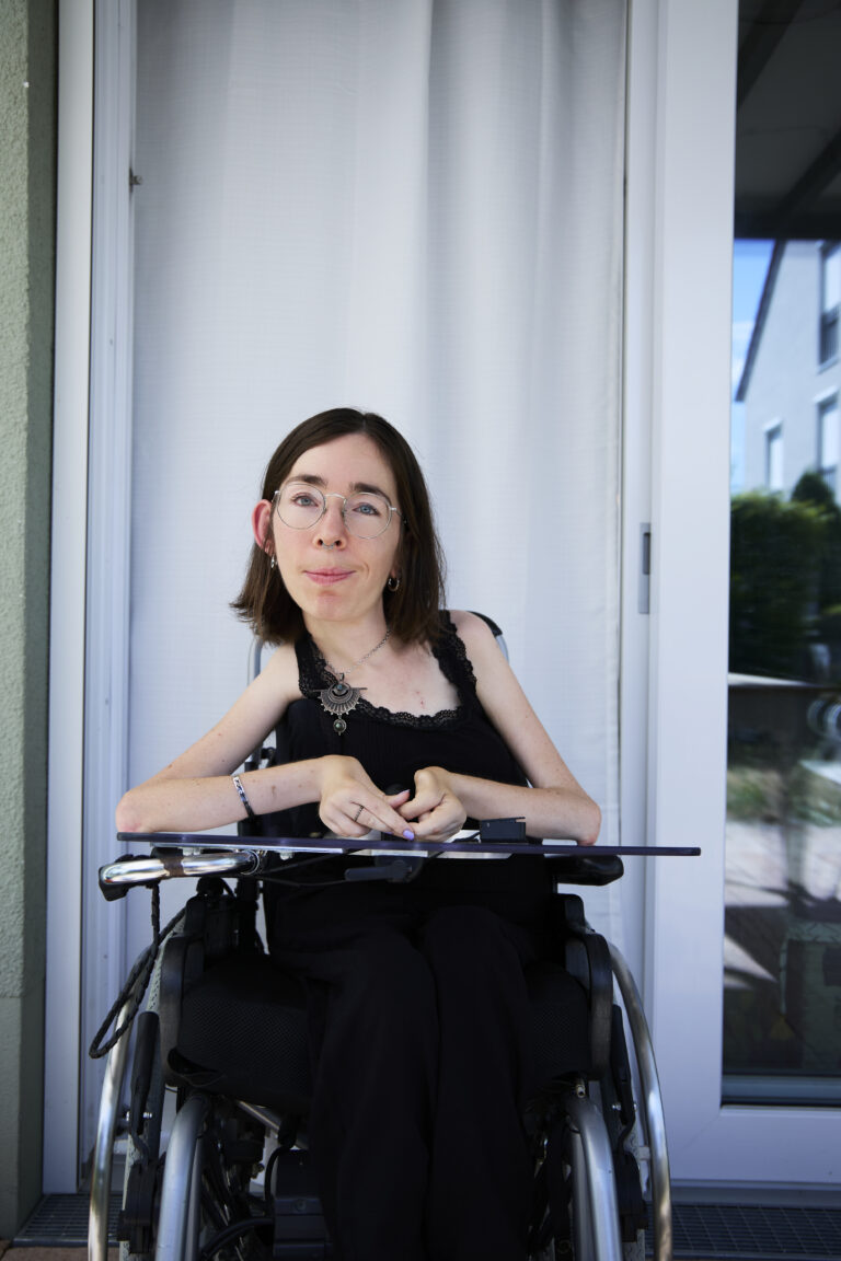 Eine Frau sitzt in einem Rollstuhl. Sie hat dunkle Haare und eine Brille. Sie schaut. mit freundlichen Blick in die Kamera.