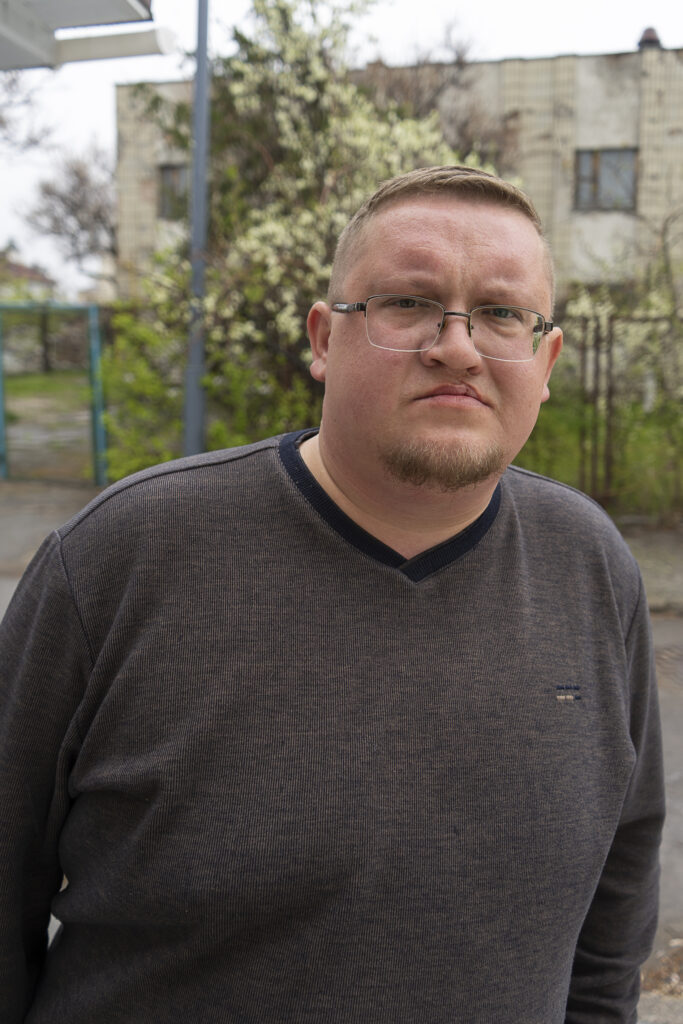 Ein Mann schaut in die Kamera. Er hat kurze blonde Haare, trägt eine Brille und ein dunkles langärmliges T-Shirt.