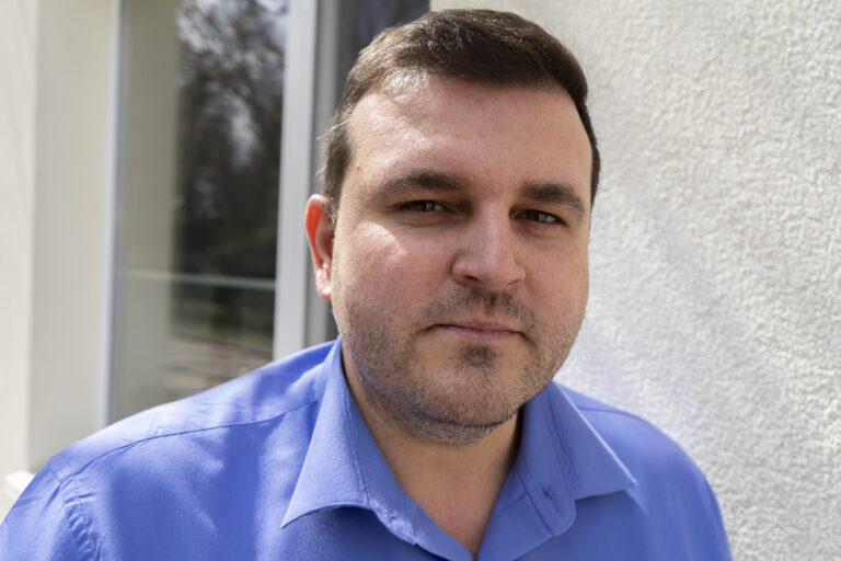 Ein Mann schaut in die Kamera. Er hat kurze braune Haare und trägt ein blaues Hemd.