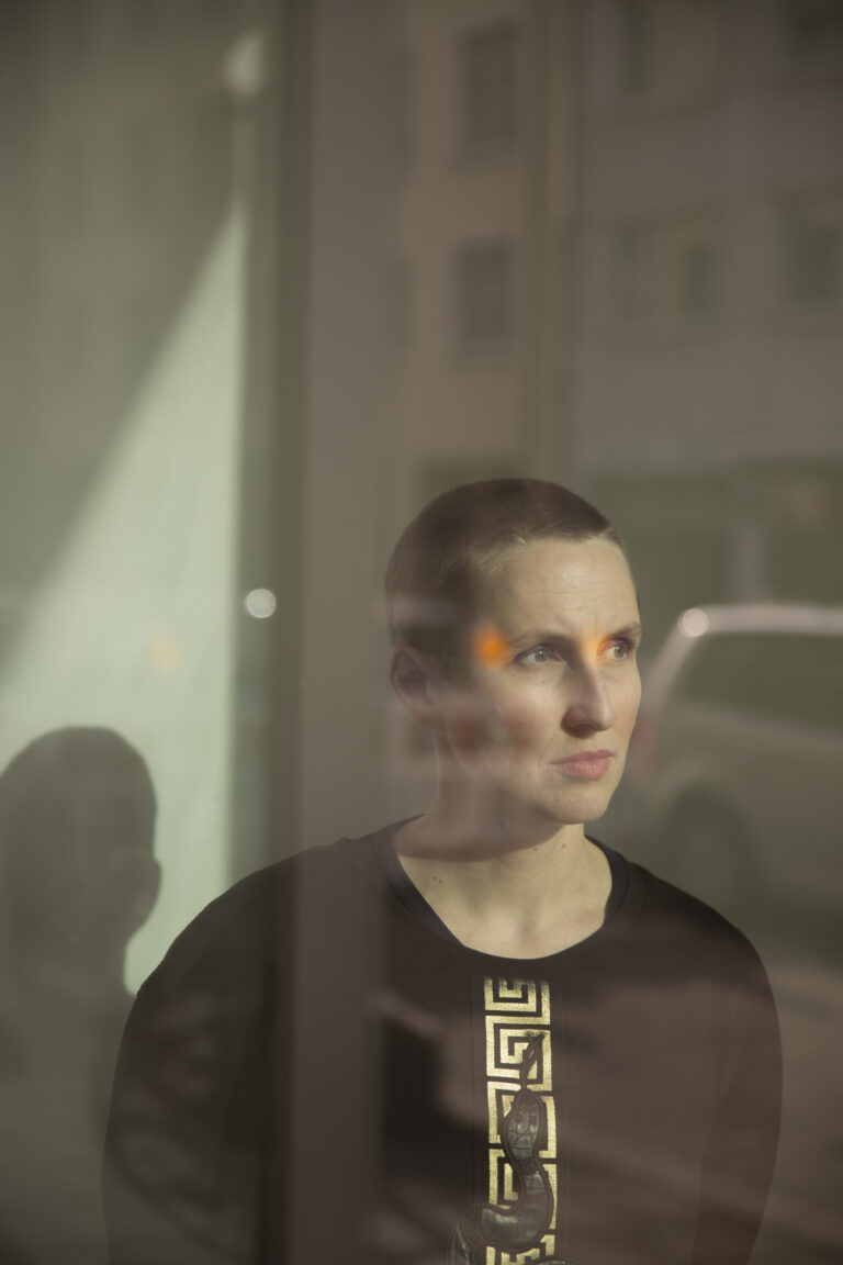 Eine junge Frau mit zusammengebunderen, braunen Haaren steht in einer Ecke. Sie trägt ein schwarzes Oberteil