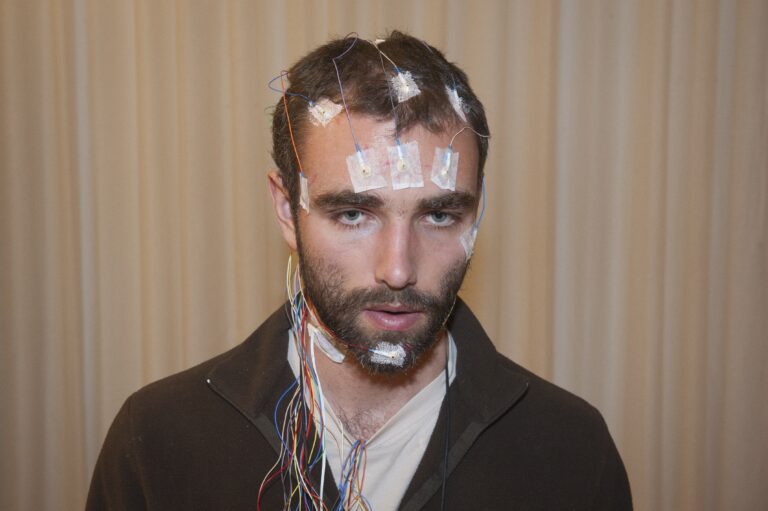 Ein Mann schaut geradeaus. Er hat braune Haare und einen braunen Bart. Am Kopf, sowie auf der Stirn und am Kinn hat er mehrere Pflaster, die mit Kabeln verbunden sind.
