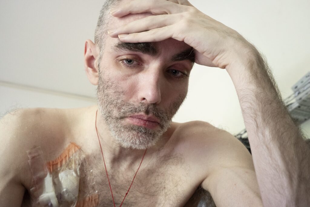 Ein Mann schaut mit ernstem Blick an der Kamera vorbei. Seine Augen sind glasig. Er stütz mit der Hand den Kopf ab. Auf der nackten Brust kleben Pflaster.