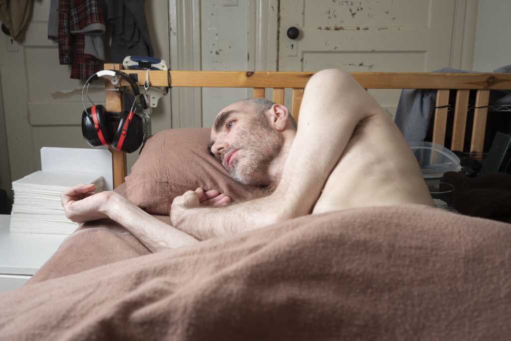 Ein Mann liegt in einem Bett. Er schaut mit starren Blick nach links. Er hat eine Glatze, blaue Augen und buschige Augenbrauen. Sein Körper ist bis zur Brust mit einer braunen Decke bedeckt.